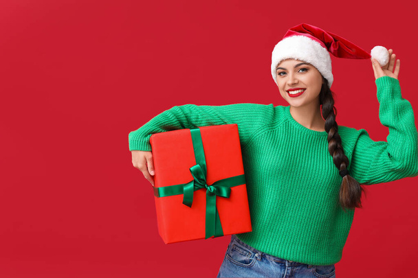 Belle jeune femme en chapeau de Père Noël avec cadeau de Noël sur fond rouge - Photo, image