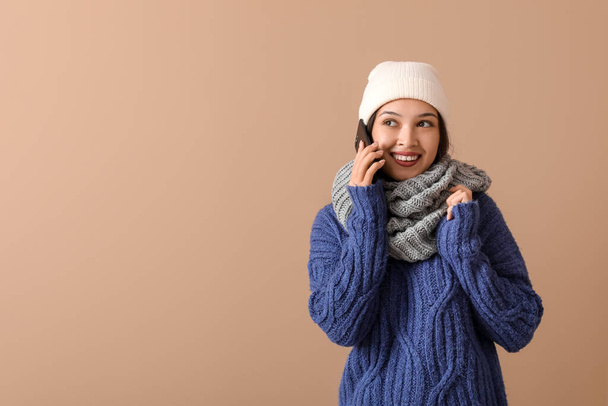 Beautiful young Asian woman in warm scarf talking by mobile phone on brown background - Photo, Image