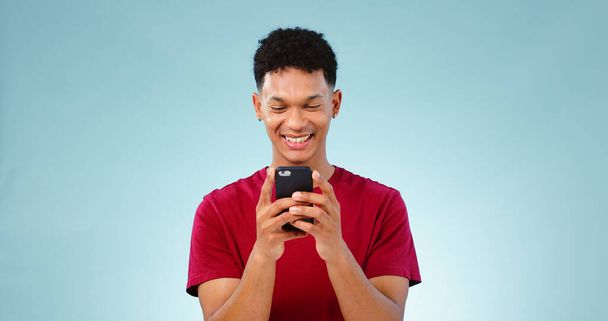 Hombre, sonrisa y mensajes de texto en el teléfono en el estudio para la comunicación se burlan de fondo azul. Joven, persona, modelo y Ciudad del Cabo con aplicación móvil, mensaje o redes sociales por internet, conexión o celular. - Foto, Imagen