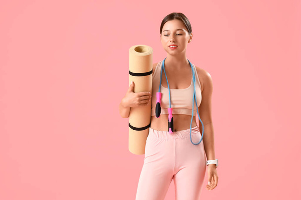 Sporty young woman with jumping rope and yoga mat on pink background - Photo, Image