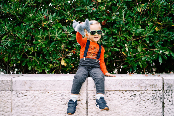 Niña en gafas de sol con un juguete suave en la mano se sienta en una valla de piedra cerca de un arbusto verde. Foto de alta calidad - Foto, imagen