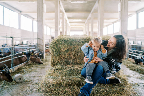 Mamá sonriente con una niña en su regazo sentada en un pajar cerca de corrales de cabra en la granja. Foto de alta calidad - Foto, Imagen