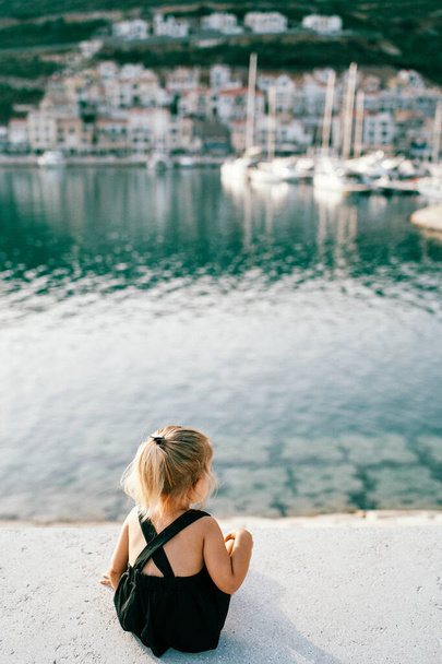 Mała dziewczynka siedzi na molo i patrzy na zacumowane jachty. Widok z tyłu. Wysokiej jakości zdjęcie - Zdjęcie, obraz