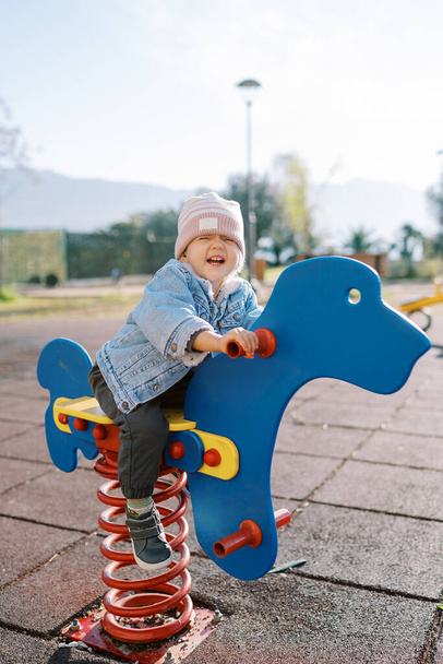 La pequeña niña riendo se balancea en un columpio en el patio de recreo sosteniendo las manijas. Foto de alta calidad - Foto, imagen