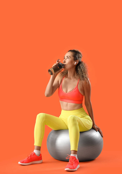 Спортивная взрослая женщина пьет воду из бутылки, сидя на фитнес-мяч на оранжевом фоне - Фото, изображение