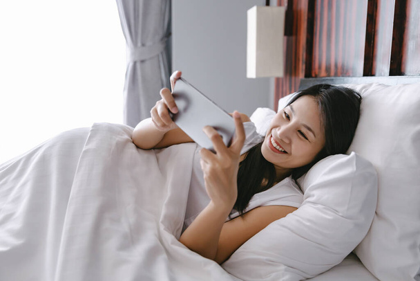 bonita mulher asiática feliz usando telefone celular na cama depois de acordar no quarto moderno em casa - Foto, Imagem