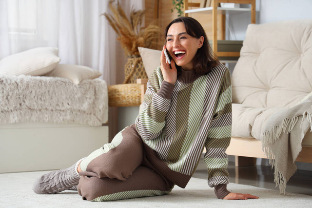 Νεαρή γυναίκα με ζεστό πουλόβερ μιλάει από το κινητό τηλέφωνο στο σπίτι - Φωτογραφία, εικόνα