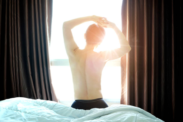 Uuden päivän aamuna herätyskello herättää huoneessa istuvan miehen. Nainen venyttää lihaksia ikkunassa. Terveydenhuollon käsitteet - Valokuva, kuva