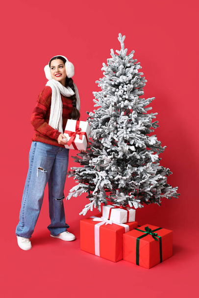 Όμορφη νεαρή γυναίκα με κουτιά δώρων κοντά στο χριστουγεννιάτικο δέντρο σε κόκκινο φόντο - Φωτογραφία, εικόνα