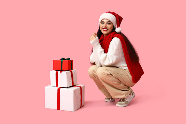 Beautiful young woman in Santa hat with Christmas gifts on pink background - Photo, Image