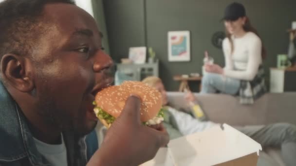 Side view medium closeup of African American guy taking bite of delicious saucy burger while chilling with company of diverse friends at home on weekend - Footage, Video