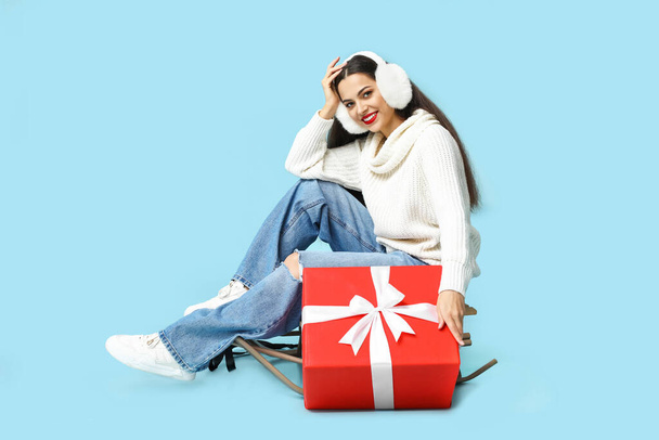 Beautiful young woman with sled and Christmas gift on blue background - Photo, Image