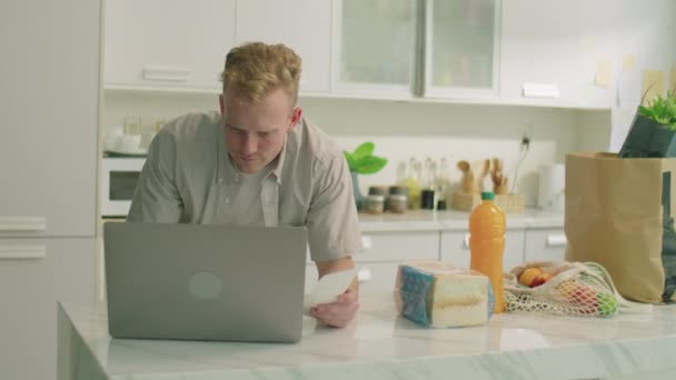 Μέτριο πλάνο του νεαρού άνδρα ακουμπά στο τραπέζι της κουζίνας με παντοπωλεία, ανάγνωση της απόδειξης και τη χρήση φορητού υπολογιστή, ενώ τη διαχείριση του προσωπικού προϋπολογισμού στο σπίτι - Πλάνα, βίντεο