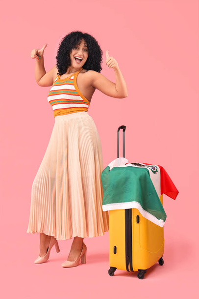 Hermosa mujer con bandera mexicana y maleta mostrando pulgares hacia arriba sobre fondo rosa - Foto, imagen
