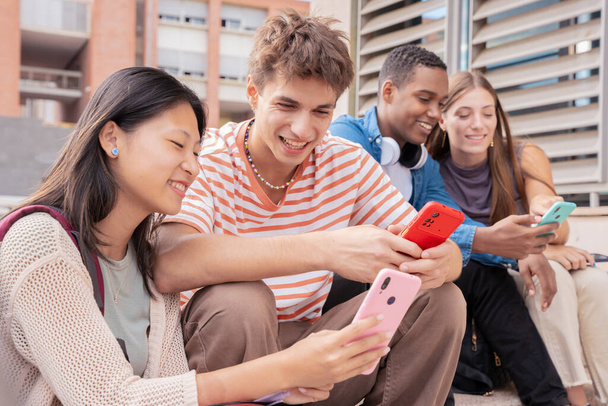 Uśmiechnięci wielorasowi studenci siedzą na zewnątrz i rozmawiają przez telefon komórkowy. Koncepcja życia uniwersytetu - Zdjęcie, obraz
