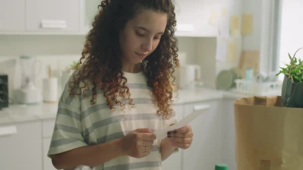 Tilt down shot of curly-haired girl standing by kitchen table with groceries on it and reading receipt from supermarket at home - Footage, Video
