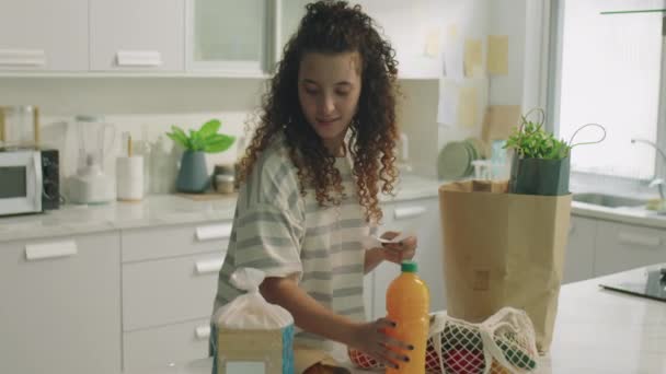 Chica joven de pie en la cocina en casa, leyendo recibo y comprobando los comestibles después de hacer compras de alimentos en el supermercado - Metraje, vídeo