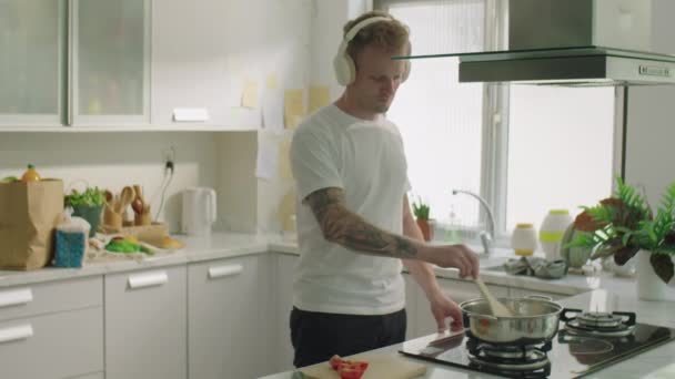 Μεσαία μακριά βολή του νέου άνδρα ακούγοντας μουσική με ασύρματα ακουστικά και χαμογελώντας ενώ το μαγείρεμα δείπνο στην κουζίνα στο σπίτι - Πλάνα, βίντεο
