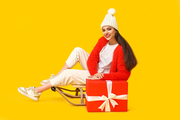 Beautiful young woman with sled and Christmas gift on yellow background - Photo, Image