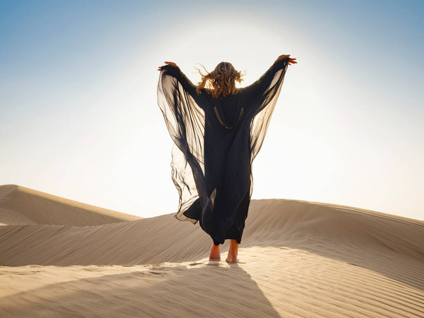 Pustynna przygoda. Młoda arabska kobieta pozująca w tradycyjnej sukni Emiratów abaya w piaskowcowych wydmach pustyni ZEA o zachodzie słońca. Rezerwat Konserwacji Pustyni w Dubaju, Zjednoczone Emiraty Arabskie. - Zdjęcie, obraz