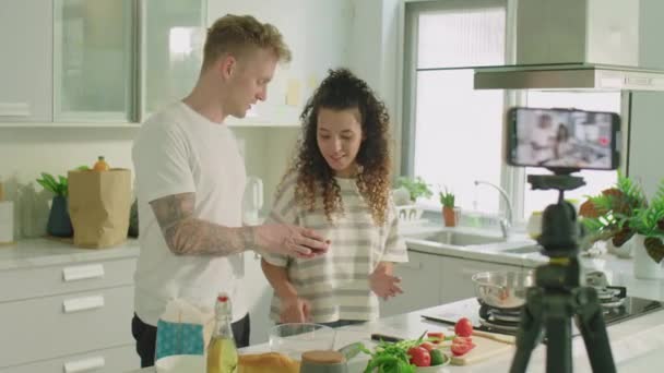Medium shot di gen Z coppia in piedi in cucina a casa e raccontare gli ingredienti alimentari durante le riprese online masterclass culinarie insieme con il telefono cellulare su treppiede - Filmati, video