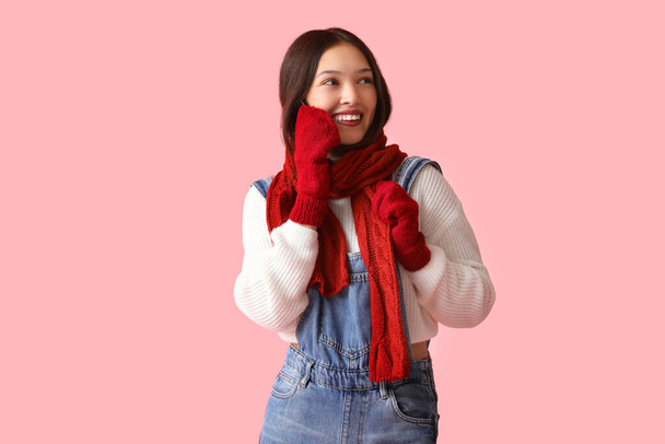 Belle jeune femme asiatique en écharpe chaude et gants parlant par téléphone mobile sur fond rose - Photo, image