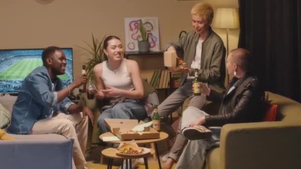 Mediana toma de cuatro alegres jóvenes diversos charlando, bebiendo cerveza y refrescos y comiendo comida rápida mientras se reúnen en el apartamento en la noche de fin de semana - Imágenes, Vídeo
