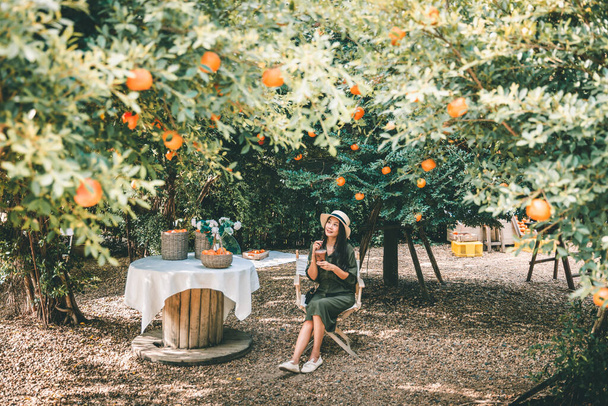 Feliz viajero asiático mujer con hielo café taza de plástico de la mano relajarse y viajar en naranja jardín café en Tailandia - Foto, Imagen