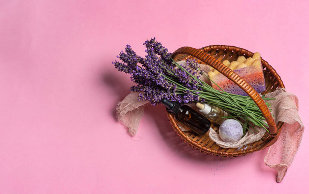Verse lavendelbloemen, etherische olie in flessen, lavendel badzout en badspons in een kleine rieten mand op een pastelroze achtergrond. met kopieerruimte - Foto, afbeelding