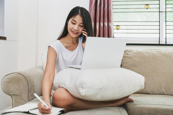 Praca z domu koncepcja, Szczęśliwa Azjatka za pomocą telefonu komórkowego i laptopa z notebooka na kanapie w domu - Zdjęcie, obraz