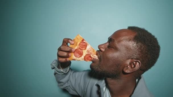 Vista lateral pecho hasta de hambre afroamericano chico comer pepperoni pizza mordida por mordida de pie sobre fondo de estudio de color turquesa - Imágenes, Vídeo