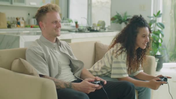 Gen Z пара сидить на дивані у вітальні, посміхається і використовує контролери, граючи відеоігри разом під час дозвілля вдома - Кадри, відео