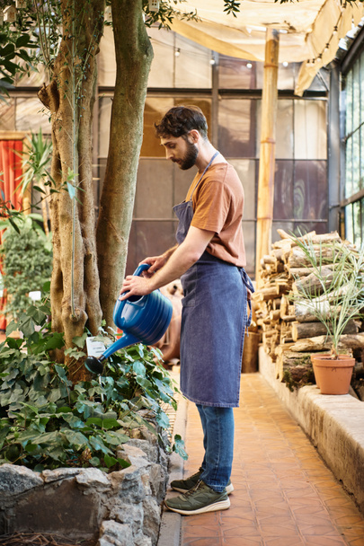 jóképű és szakállas kertész farmer kötény öntözés növények üvegház, kertészet koncepció - Fotó, kép