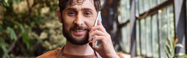 hermoso jardinero barbudo en delantal sonriendo y hablando en el teléfono inteligente en invernadero, pancarta - Foto, Imagen