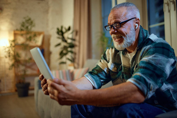 Side view portrait of mature man in glasses sitting on couch in living room and watching movie in tablet and in headphones on head. Concept of love, retirement life, pensioners, cozy, winter holidays. - Photo, Image