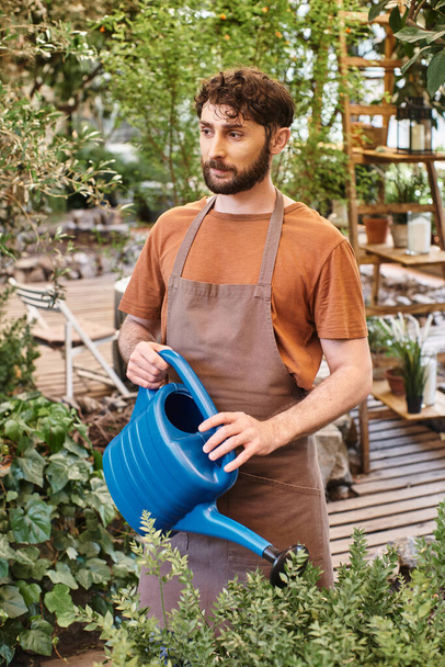 professionele tuinman in linnen schort besproeiing groene struik met blauwe gieter in kas - Foto, afbeelding