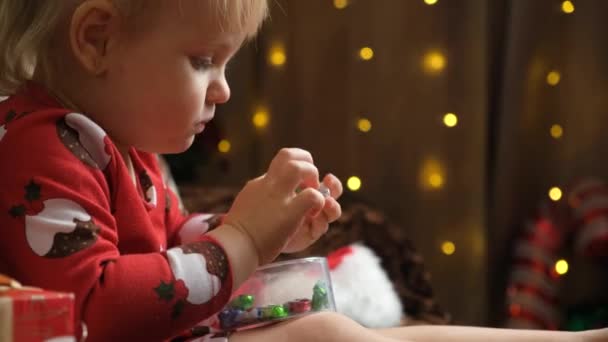 Pieni kaksivuotias tyttö syö karkkia jouluna istuessaan kotona lahjojen joukossa.. - Materiaali, video