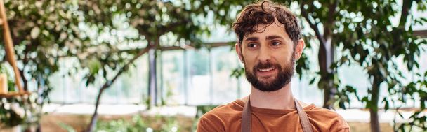professional headshots, cheerful bearded gardener in apron and gloves posing in greenhouse, banner - Photo, Image