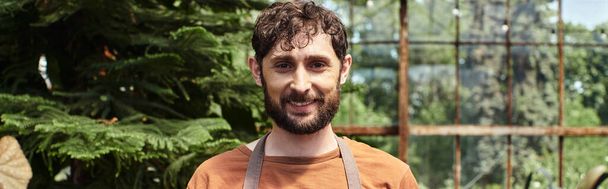 happy bearded gardener looking at camera around green plants in greenhouse, horticulture concept - Photo, Image