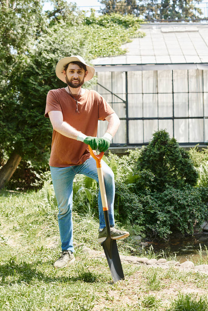 веселий фермер з бородою в сонячному капелюсі і стоїть з лопатою біля рослин і теплиці - Фото, зображення