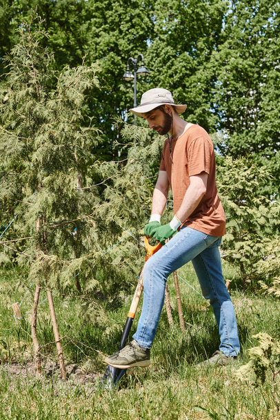 веселый фермер с бородой в шляпе от солнца и копать лопатой рядом с растениями и теплицей - Фото, изображение