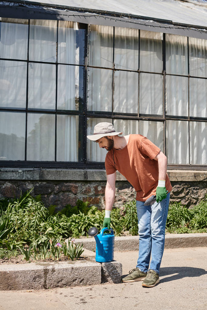pohledný a vousatý zahradník ve slunečním klobouku při zalévání plechovky a držení lopaty na venkově - Fotografie, Obrázek