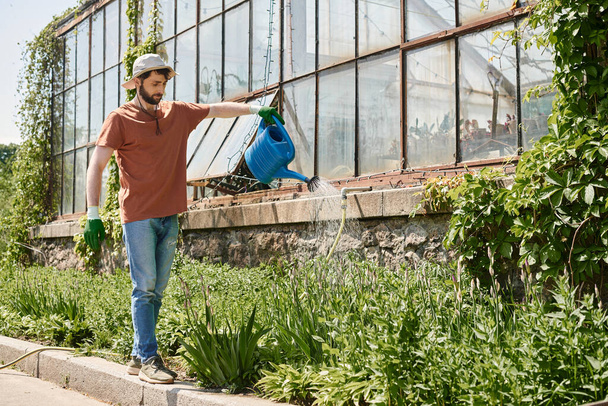handsome and bearded gardener in sun hat watering plants near greenhouse in countryside - Photo, Image