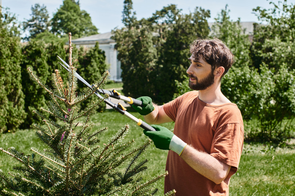 бородатый садовник в перчатках обрезает ель большими садовыми ножницами во время работы на открытом воздухе - Фото, изображение