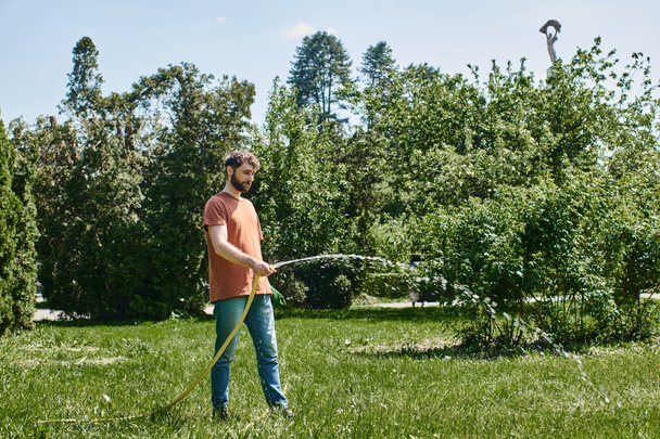 бородатый садовник в повседневной одежде, держащий перчатки и поливающий зеленую лужайку во время работы на открытом воздухе - Фото, изображение