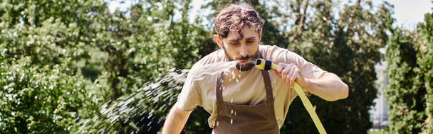 vousatý zahradník v plátnové zástěře pitná voda z hadice po práci v zahradě, otevřený prapor - Fotografie, Obrázek