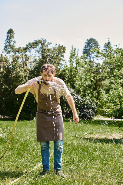 bearded gardener in linen apron drinking water from hose after working in garden, candid photo - Photo, Image