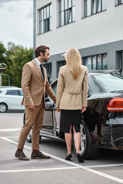 brodaty elegancki mężczyzna otwarcie tylne drzwi luksusowego samochodu w pobliżu modnej blondynki businesswoman na ulicy miasta - Zdjęcie, obraz