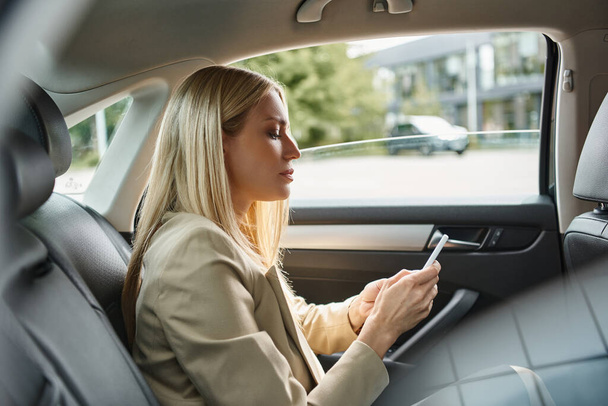 Вид сбоку блондинки-бизнесвумен в формальной одежде и с мобильным телефоном в роскошном автомобиле на улице - Фото, изображение
