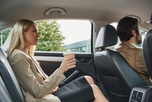 blonde businesswoman holding coffee to go near elegant man driving luxury car on urban street - Photo, Image